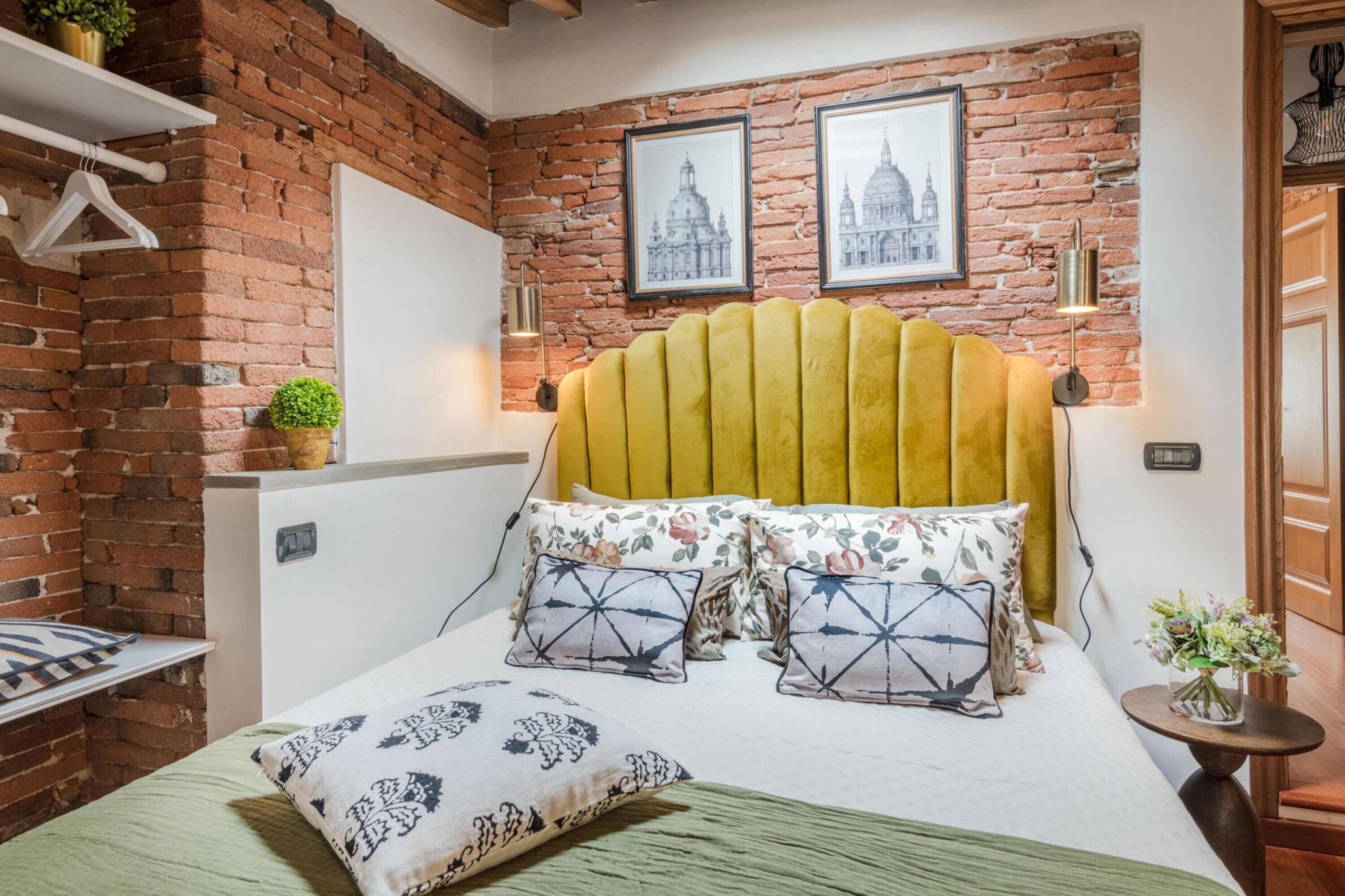 Ein Zimmer mit einer Ziegelwand, einem gelben, gepolsterten Kopfteil, floralen und geometrischen Kissen sowie gerahmten Bildern von Gebäuden.