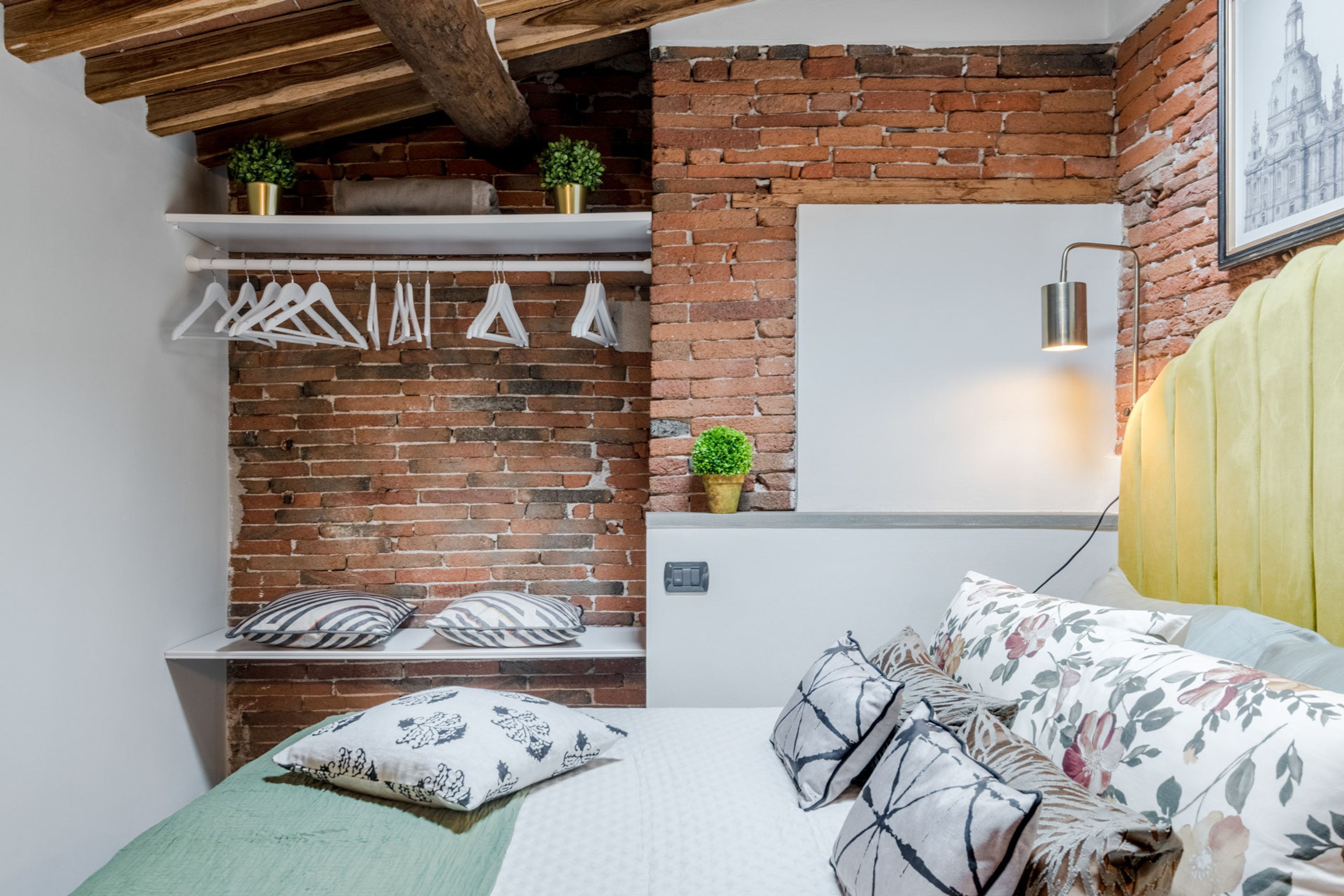 Une chambre avec un mur en briques, un lit recouvert d’une literie à motifs floraux, un plaid vert et un luminaire suspendu.