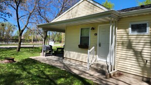 A single-story house with a covered porch, a grill, and a small window. - Studio Cottage at Lake Road Campground, on the beautiful Mississippi River. (Trempealeau)