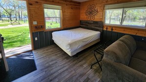 A wooden cabin room with a bed, a couch, and two windows offering views of a park. - Studio Cottage at Lake Road Campground, on the beautiful Mississippi River. (Trempealeau)