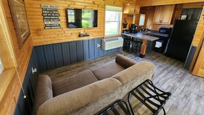 A cozy cabin interior with a wooden interior, a sofa, a TV mounted on the wall, a kitchen area with a refrigerator, and a sign listing activities like 'Go barefoot' and 'Read a book'. - Studio Cottage at Lake Road Campground, on the beautiful Mississippi River. (Trempealeau)