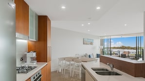A modern kitchen with a built-in stove, a coffee machine, and a dishwasher. There is a dining area with a table and chairs. The kitchen has a large window with a view of the outdoors. - Boasting coveted north facing views and an unrivalled coastal lifestyle. (Tweed)