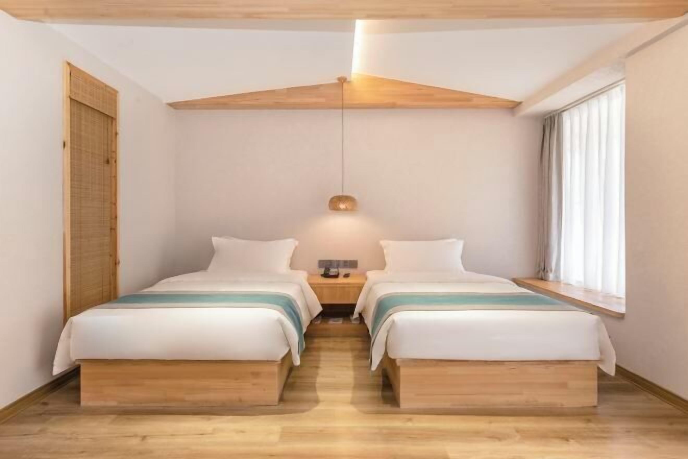 A bedroom with two beds, wooden flooring, a hanging light fixture, and a window with curtains.