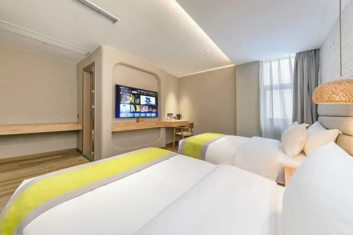 A hotel room with two beds, a TV mounted on the wall, a desk, and a window with curtains.