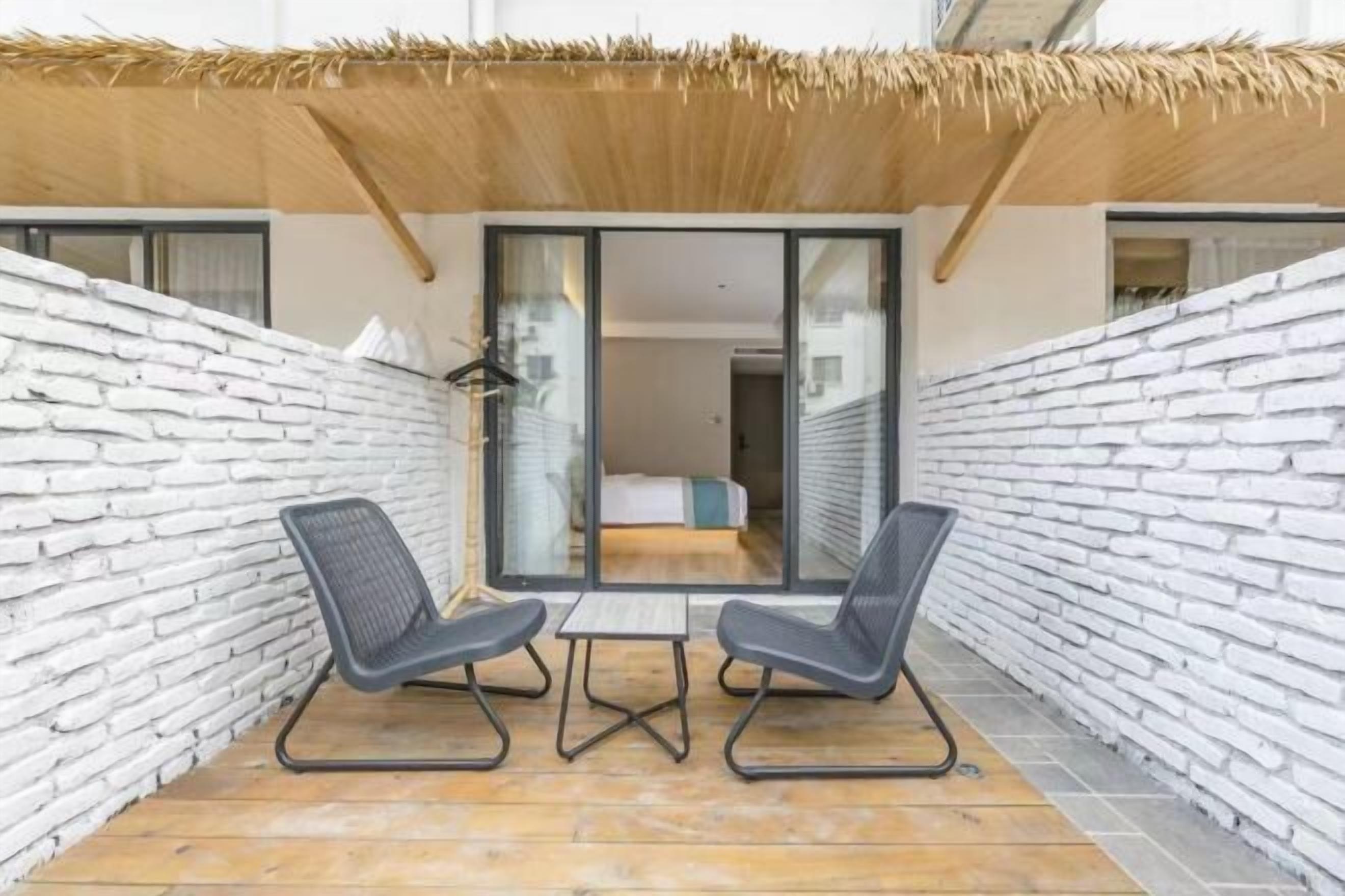 Un patio con dos sillones de descanso y una pequeña mesa, techo de paja y pared de ladrillo blanco.