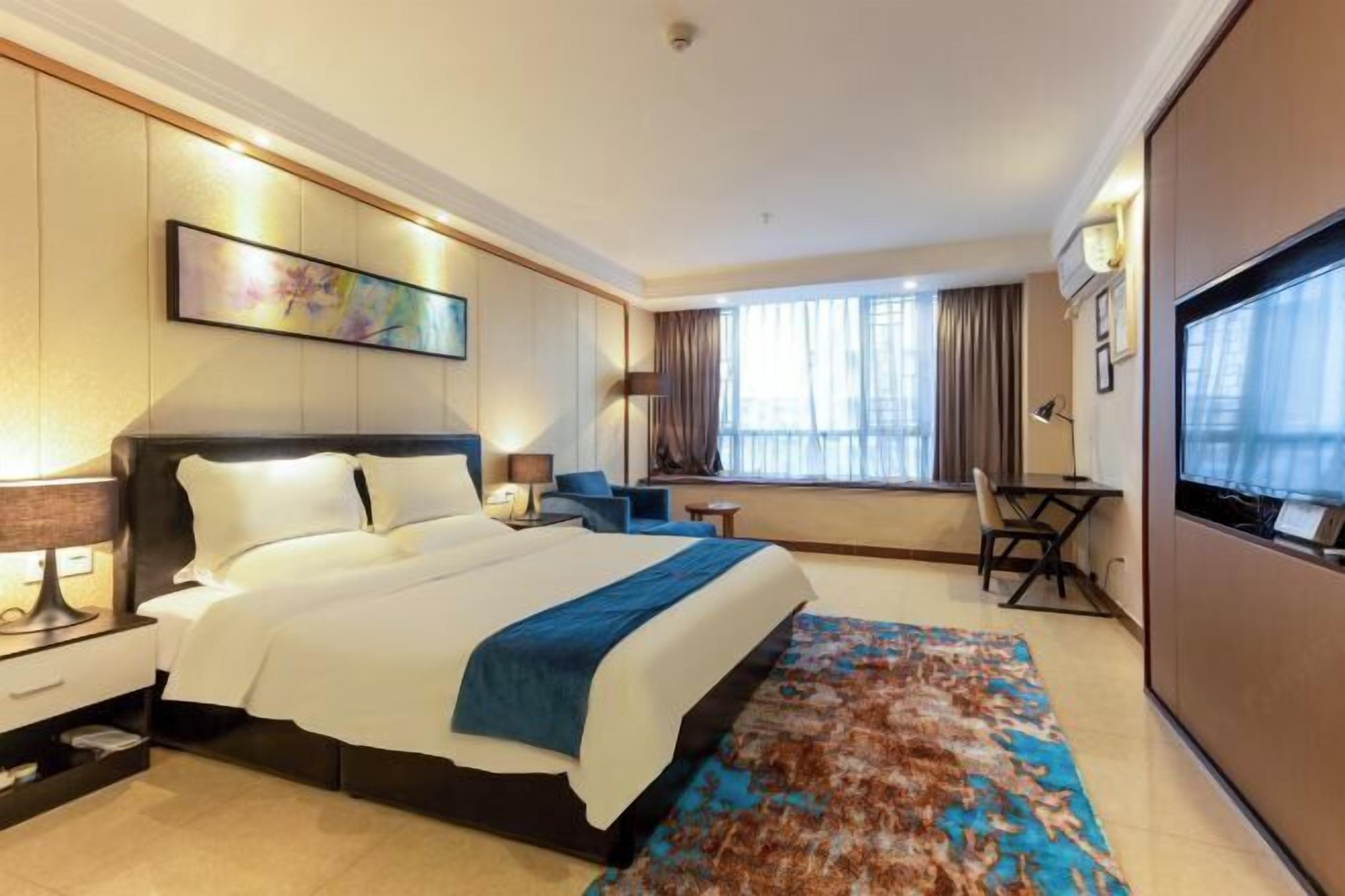 A hotel room with a large bed, a desk, a chair, a TV, and a window with curtains.