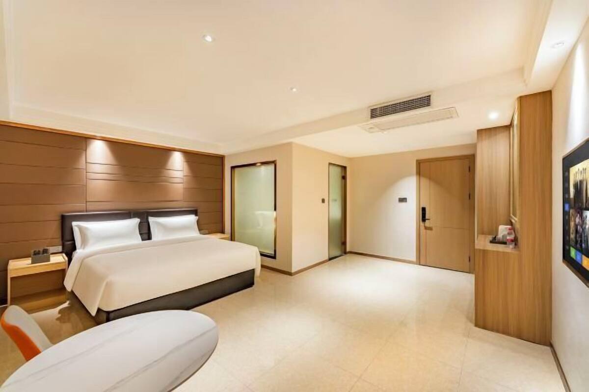A modern hotel room with a large bed, a bedside table, a chair, a wardrobe, and a door leading to another room.