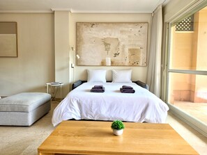 A hotel room with a large bed, a small side table, a grey sofa, a wooden table with a plant, and a large window. - SR Dunas de Carib Playa Marbella (Marbella)