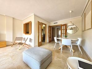 A modern living room with a grey ottoman, a dining table with white chairs, a wooden chair with a woven backrest, and a large mirror on the wall. - SR Dunas de Carib Playa Marbella (Marbella)