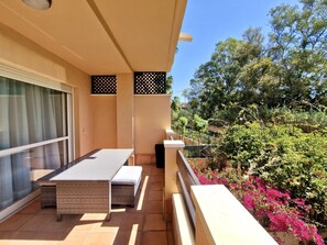 Un balcón con mesa y sillas, una puerta corredera de vidrio y vistas a plantas y flores.