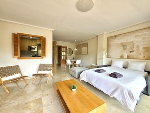 A hotel room with a bed, a wooden bench, a small table with a plant, a desk with chairs, and a kitchen area visible through a window.