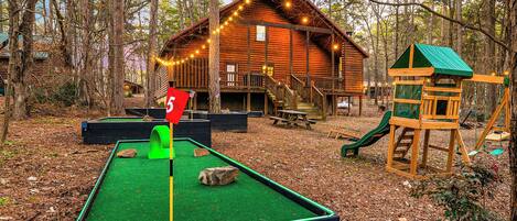 A wooden cabin with a deck and string lights, surrounded by trees.