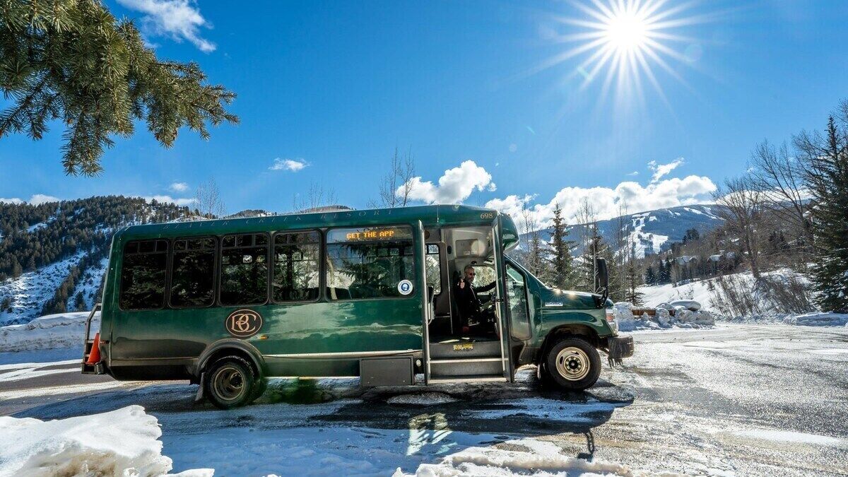 A green bus with a sign that reads 'GET THE APP' parked on a snowy road with mountains in the background.