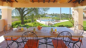 Un patio al aire libre con una mesa preparada para dos personas, rodeada de sillas, con vistas a una piscina y exuberante vegetación.
