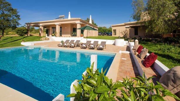Zona de piscina con sillones de descanso y una pérgola, rodeada de follaje y un edificio con techo de tejas.
