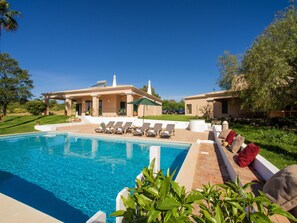 Ein Poolbereich mit Liegestühlen und einer Pergola, umgeben von Grünflächen und einem Gebäude mit Ziegeldach.