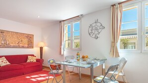 A modern living room with a red sofa, a glass dining table, and metal chairs. There is a painting on the wall and a window with curtains. - Boulevard Louis Blanc by Interhome (Saint-Tropez)