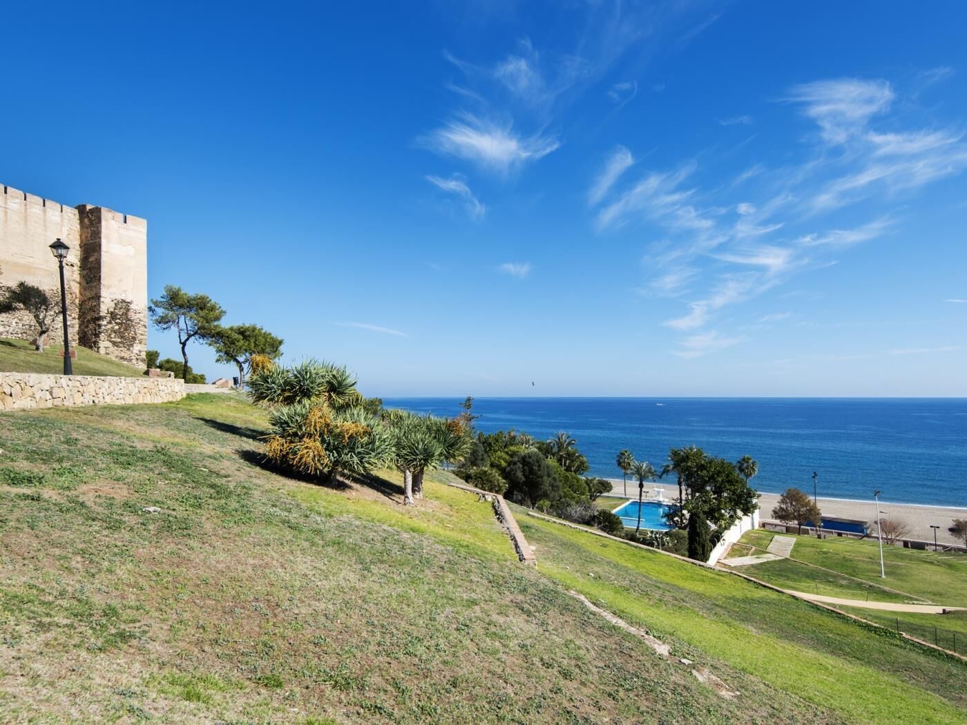 A coastal view with a stone building, a grassy slope, and a pool area.