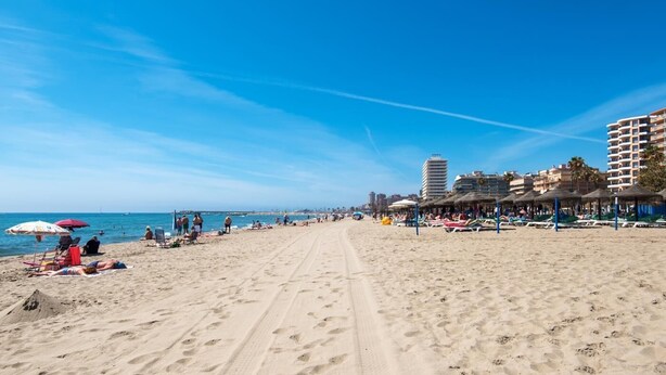 Ein Strand mit Liegestühlen und Sonnenschirmen, ein klarer Himmel und Gebäude im Hintergrund.