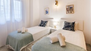 Ein Hotelzimmer mit zwei Betten, jedes mit einer grünen Decke und einem zusammengerollten Handtuch. Es gibt ein Fenster mit durchsichtigen Vorhängen, eine Wandleuchte und zwei gerahmte Bilder.