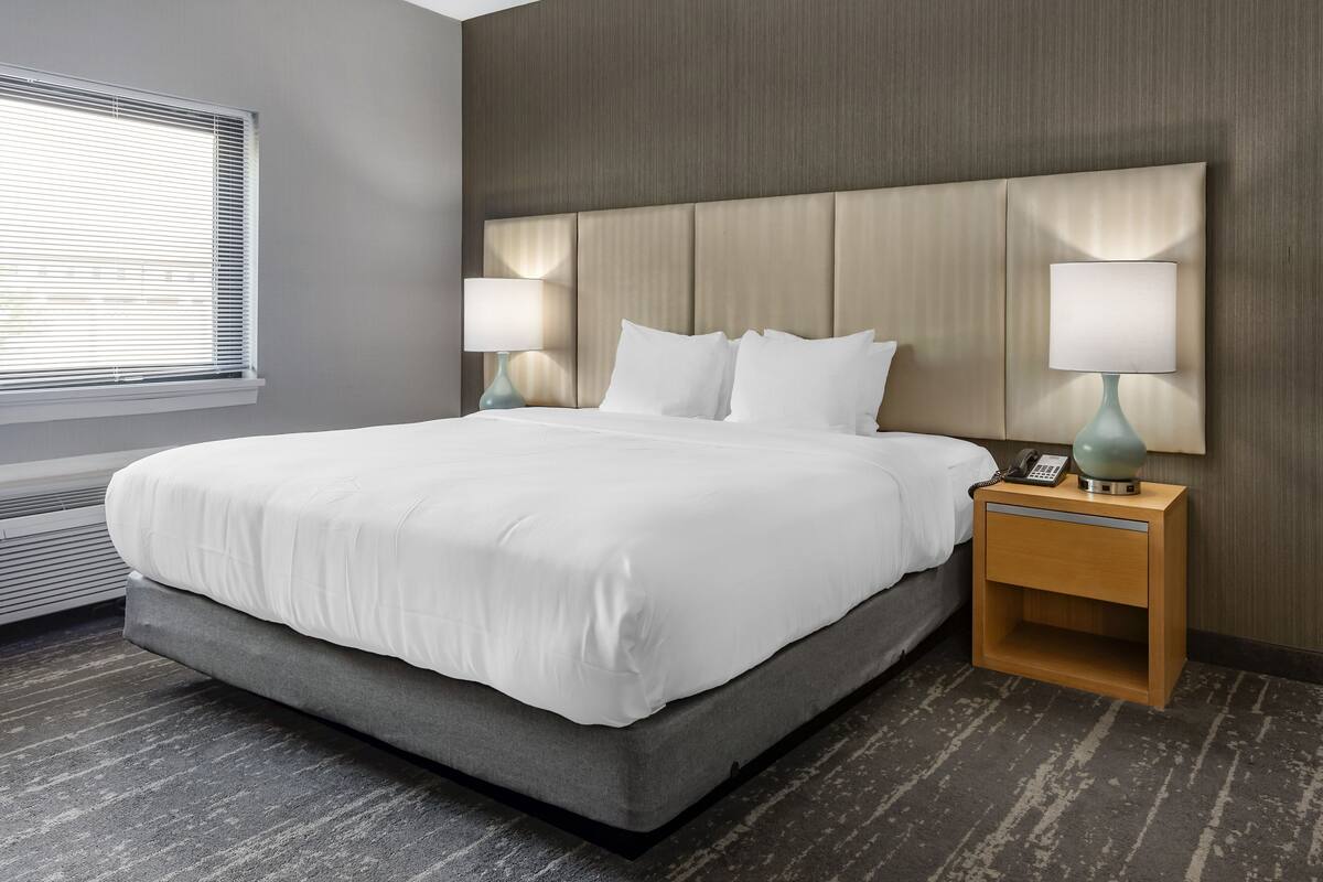 A modern hotel room with a large bed, two bedside lamps, a nightstand with a phone, and a window with blinds.