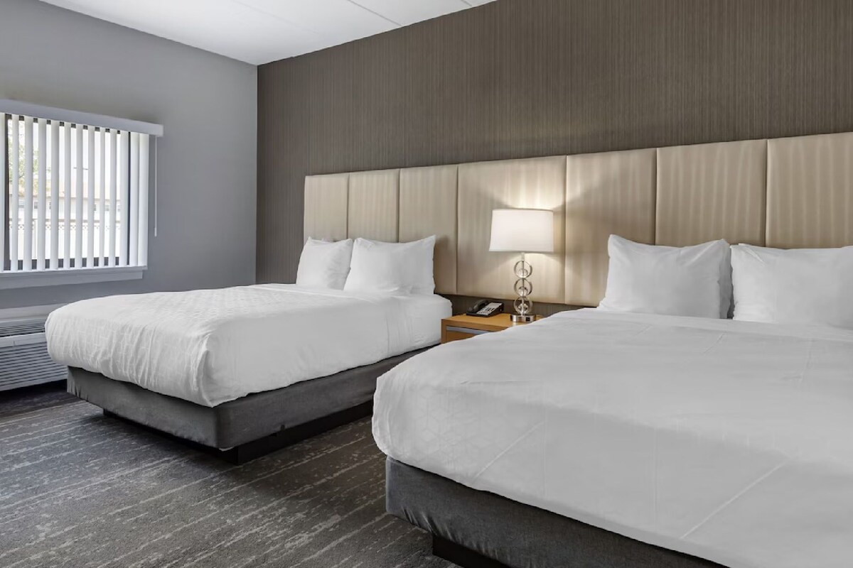 Habitación de hotel con dos camas, una mesita de noche con lámpara y una ventana con persianas.