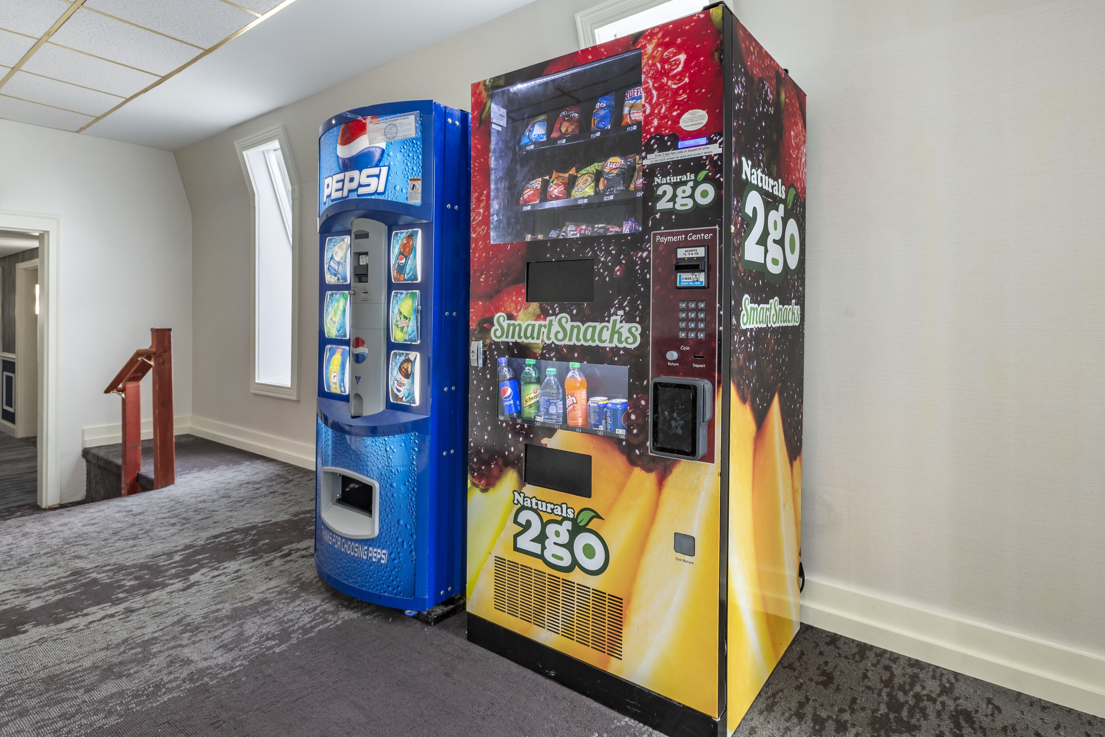 Two vending machines in a hallway with a carpeted floor and a wooden staircase.