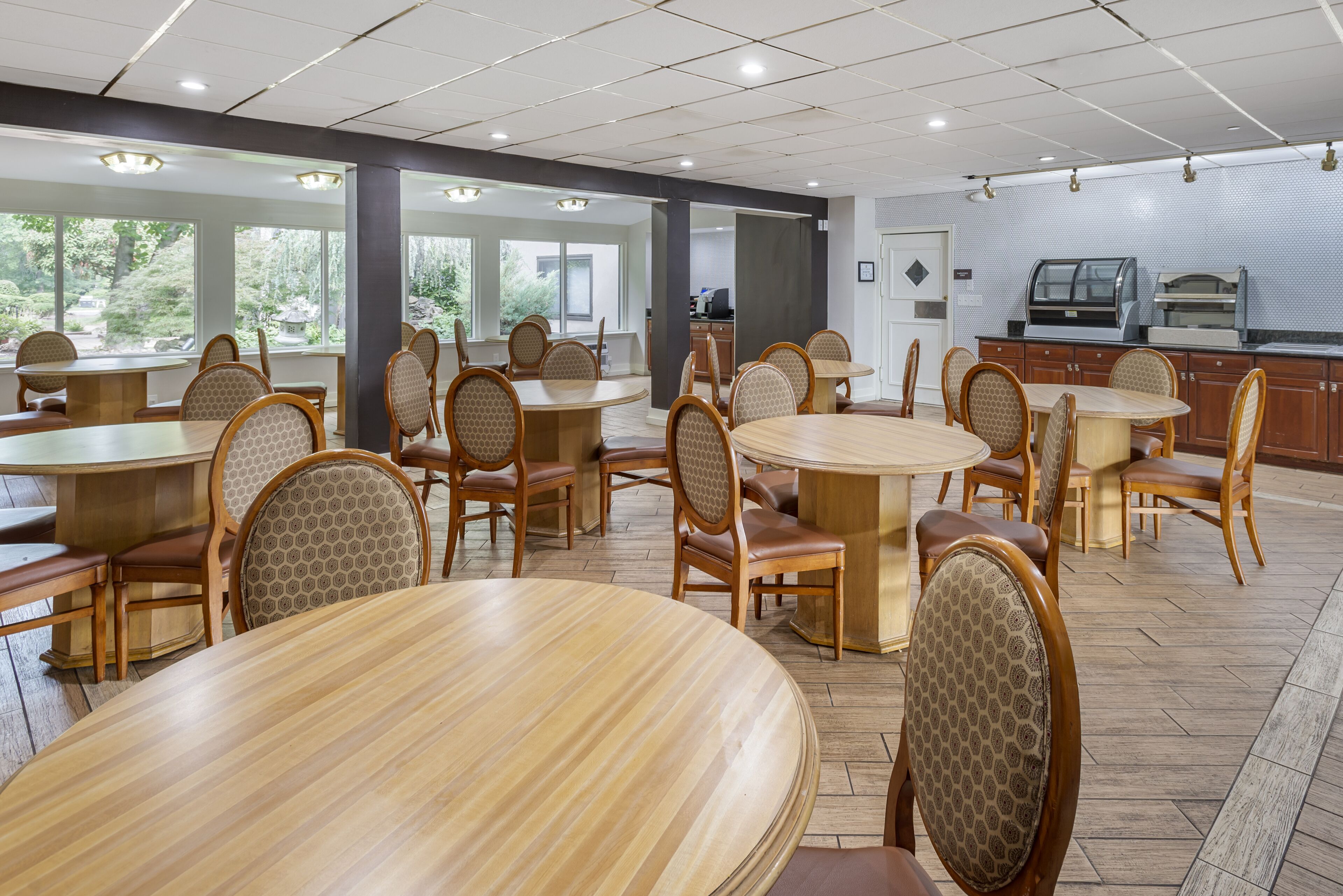 Un vaste espace dédié à la salle à manger, avec plusieurs tables et chaises rondes, de grandes fenêtres offrant une vue sur la verdure, et un coin cuisine doté d’un présentoir.