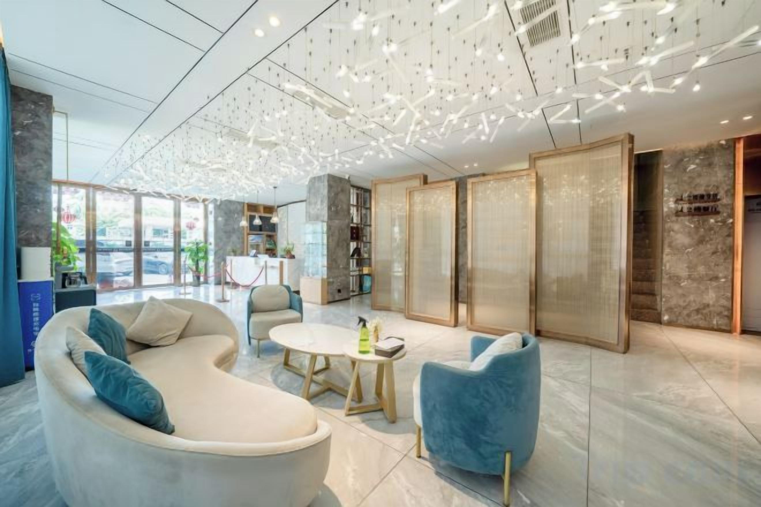 A modern lobby with a curved sofa, a round table, and blue armchairs. There are bookshelves and a large window with a view.