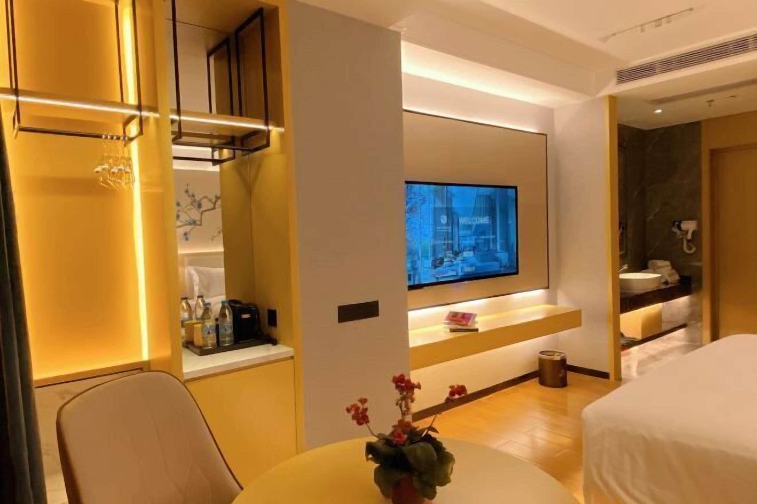 A modern hotel room with a flat-screen TV mounted on the wall, a small table with a vase of flowers, and a bed with white bedding.