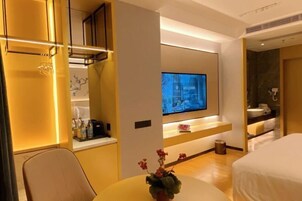 A modern hotel room with a flat-screen TV mounted on the wall, a small table with a vase of flowers, and a bed with white bedding.