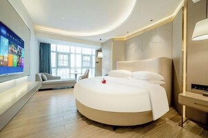 Quarto de hotel moderno com uma cama redonda, televisão presa na parede, sofá e uma janela grande.
