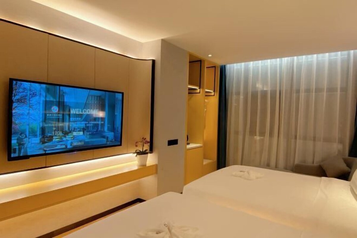 Habitació d'hotel moderna amb un televisió plana gran penjada a la paret, un llit amb roba blanca, una taula de nit i una finestra amb cortines de tul.