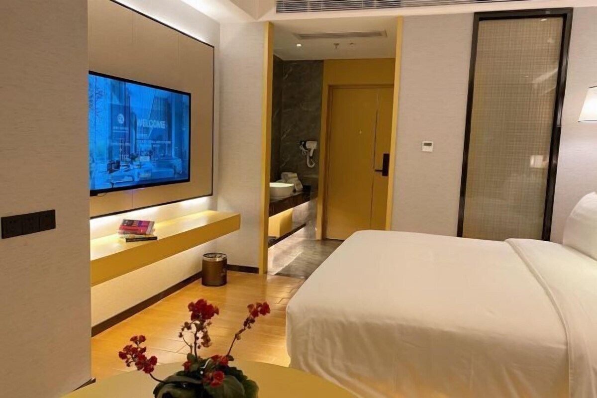 Habitació d'hotel moderna amb un llit gran, un televisor penjat a la paret, un taulell petit amb un ram de flors i un bany visible a través d'una porta oberta.