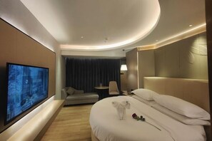 Una habitación de hotel moderna con una cama grande, un televisor de pantalla plana montado en la pared, una pequeña zona de estar y un ventanal con cortinas.