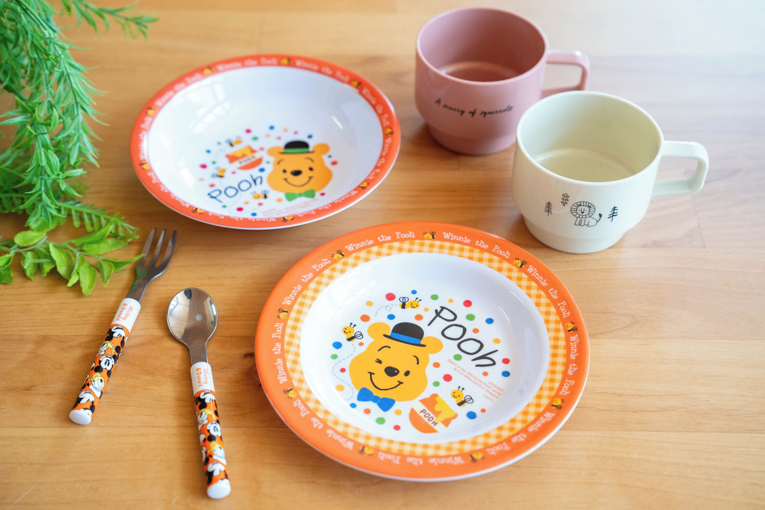 Une table avec deux assiettes à l’effigie de Winnie l’ourson, une tasse rose, une tasse blanche, une fourchette et une cuillère.