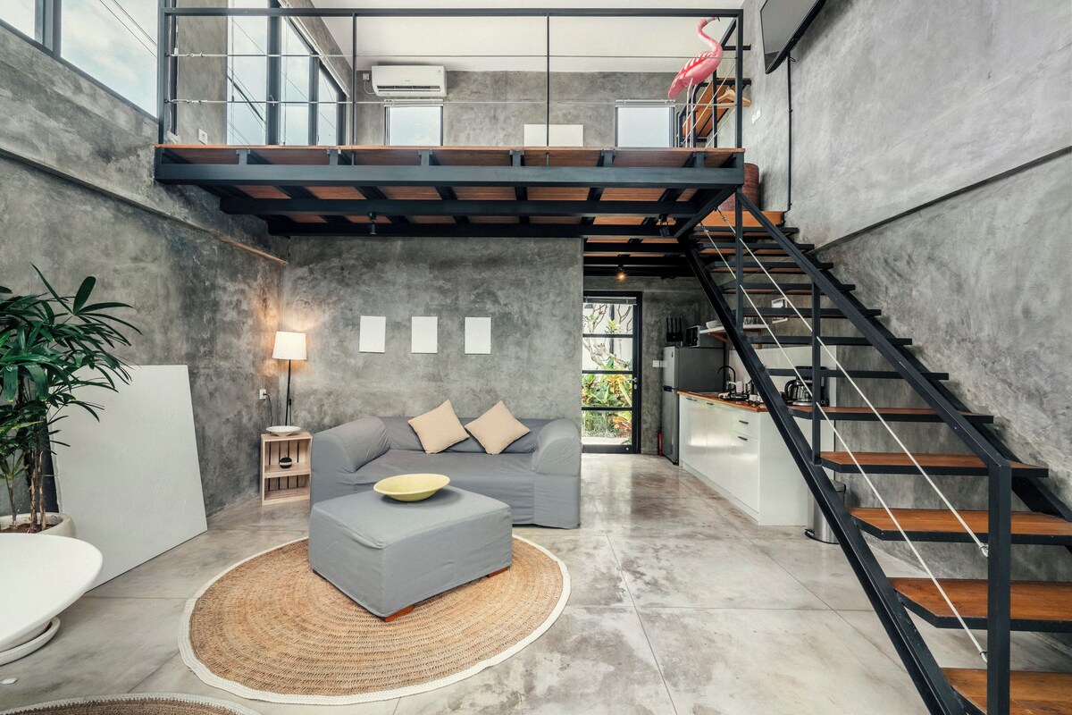 A modern loft-style living space with a grey sofa, a round coffee table, a staircase, and a large window.