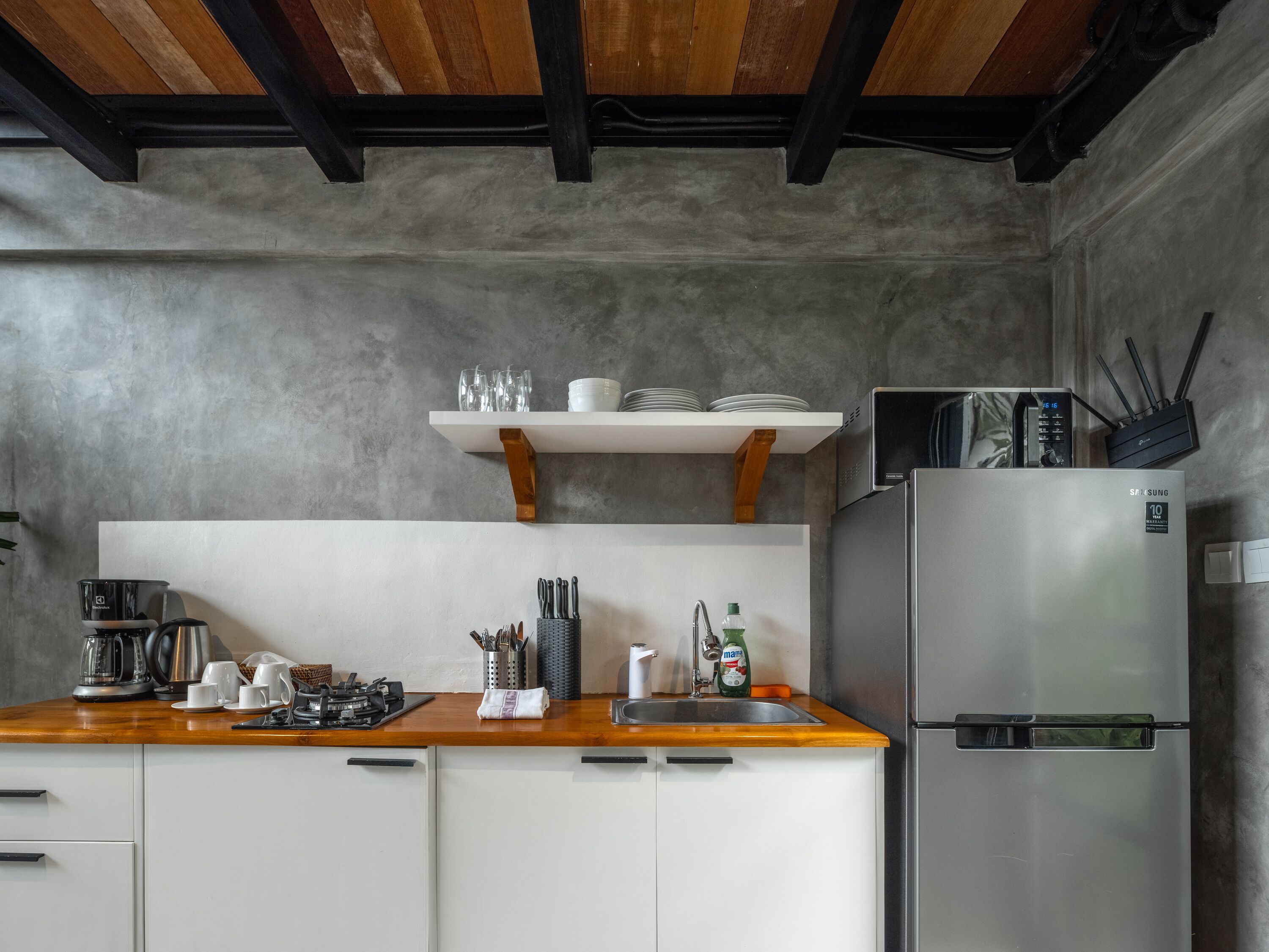 A compact kitchen with white cabinets, a stainless steel refrigerator, a wooden countertop, and a shelf above holding dishes and glasses.