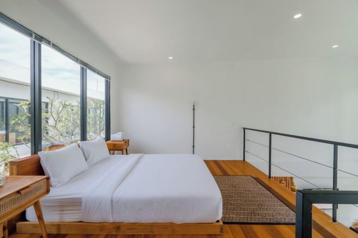 Un dormitorio moderno con una cama grande, una mesita de noche de madera, un escritorio pequeño y vistas al exterior a través de ventanas amplias.