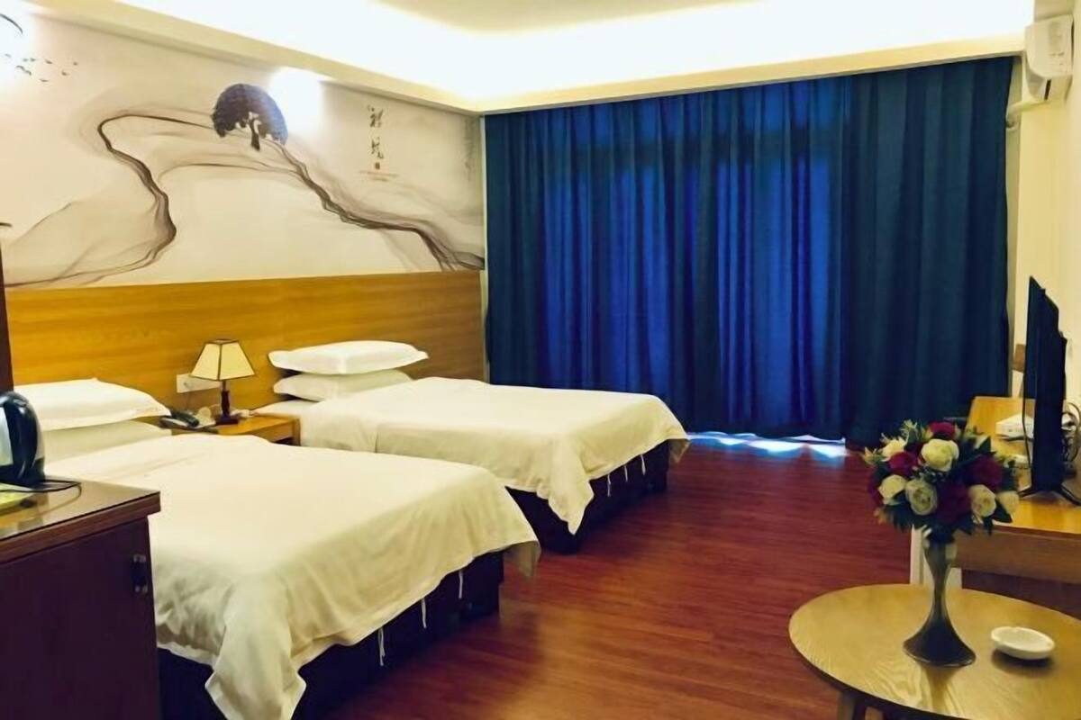 Een hotelkamer met twee bedden, een houten hoofdbord, een bureau met een lamp, een vaas met bloemen en een groot blauw gordijn.