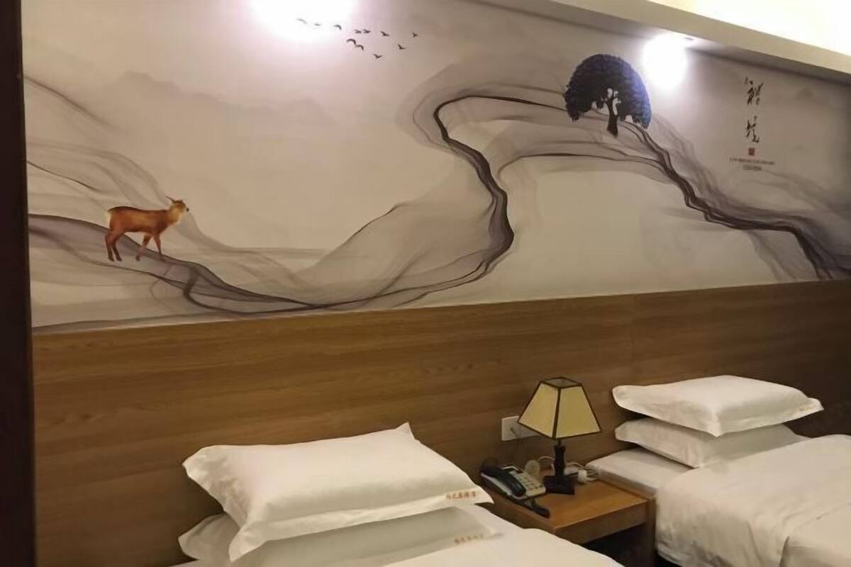 Een hotelkamer met een houten hoofdeinde, twee bedden met witte lakens, een nachtkastje met een lamp en een muurschildering van een hert en een boom.