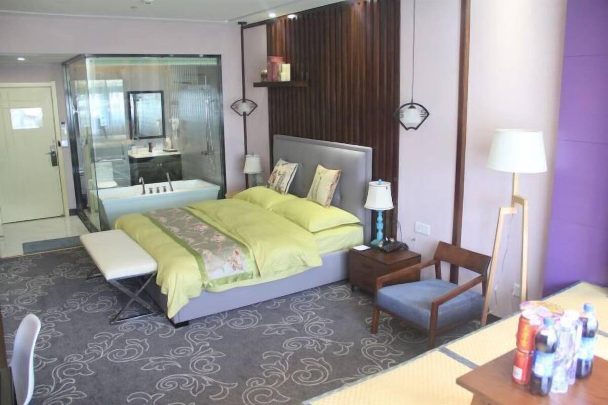 Une chambre d’hôtel comprenant un lit, des tables de chevet, une chaise, un bureau et une salle de bain séparée par une paroi en verre.