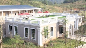 Un edificio de dos pisos con techo plano, un balcón con sombrillas rojas y un jardín.