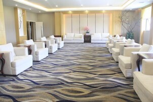 Un hall de hotel con alfombra estampada, filas de sillones blancos y una zona de recepción con un sofá y una mesita.