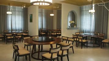 A hotel dining area with multiple round tables and chairs, large windows with curtains, and a modern chandelier.