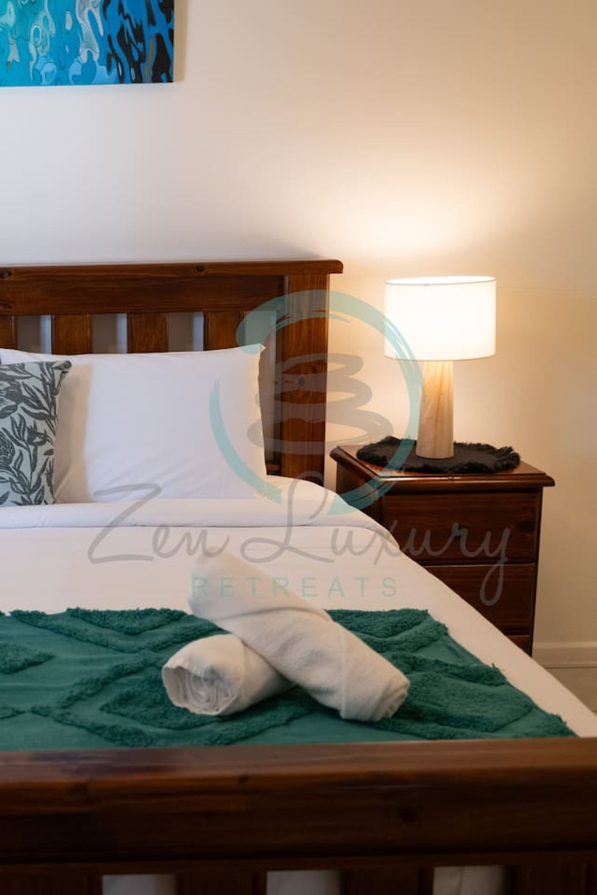 A neatly made bed with a green blanket and a rolled towel, a bedside lamp, and a nightstand with a decorative item.