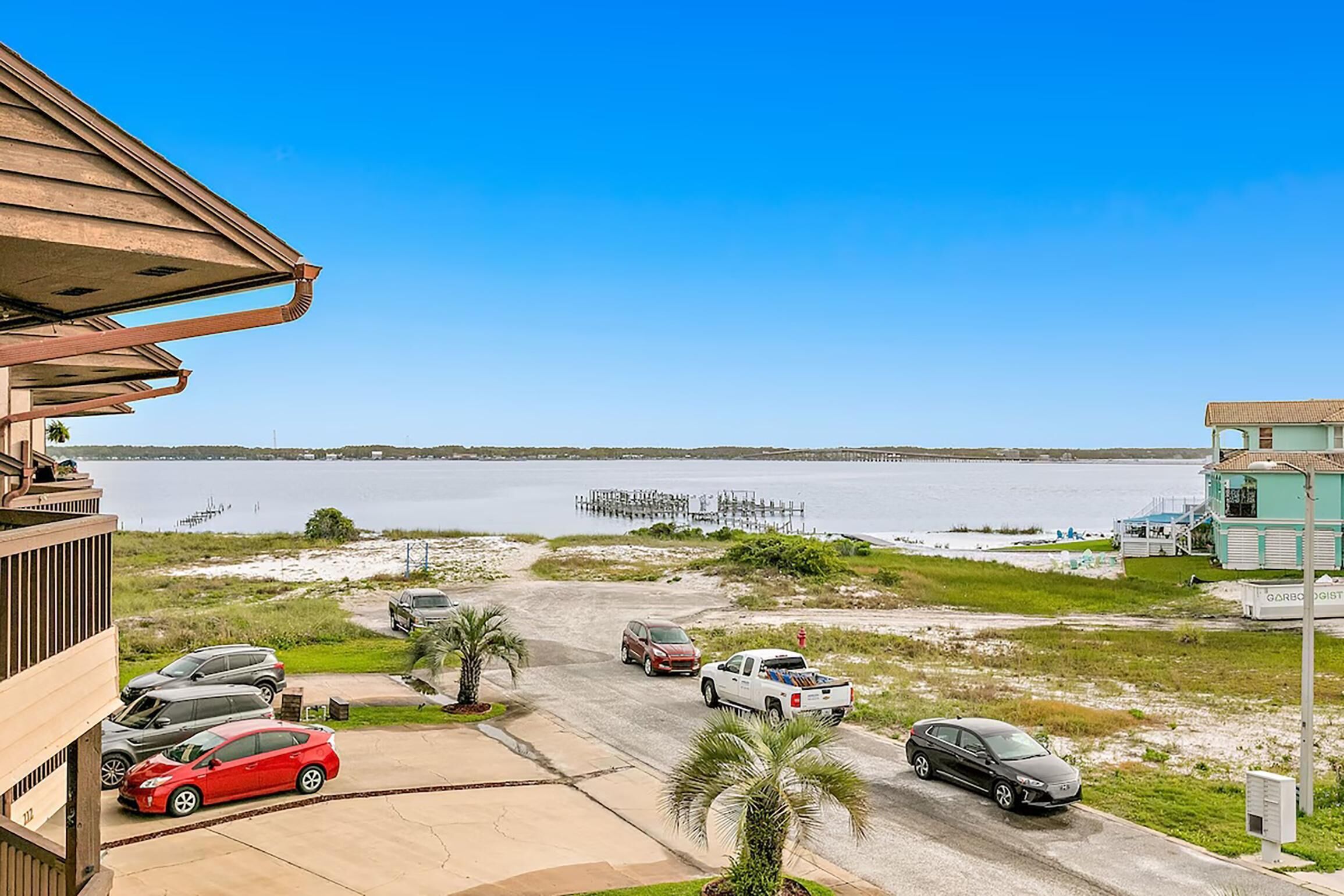 A view of a parking area with several cars, a building with a balcony, and a body of water in the background.