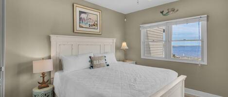 A bedroom with a large bed, a ceiling fan, a window with a view of the sea, a framed picture on the wall, and a small side table with a lamp.