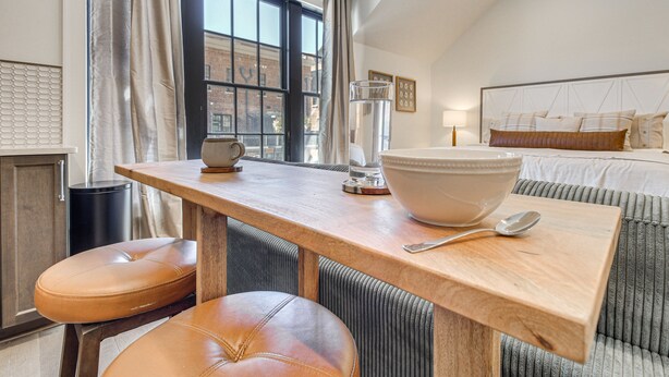 Une chambre moderne avec une grande fenêtre, une table en bois sur laquelle se trouvent un bol et une cuillère, et un lit doté d’une tête de lit.