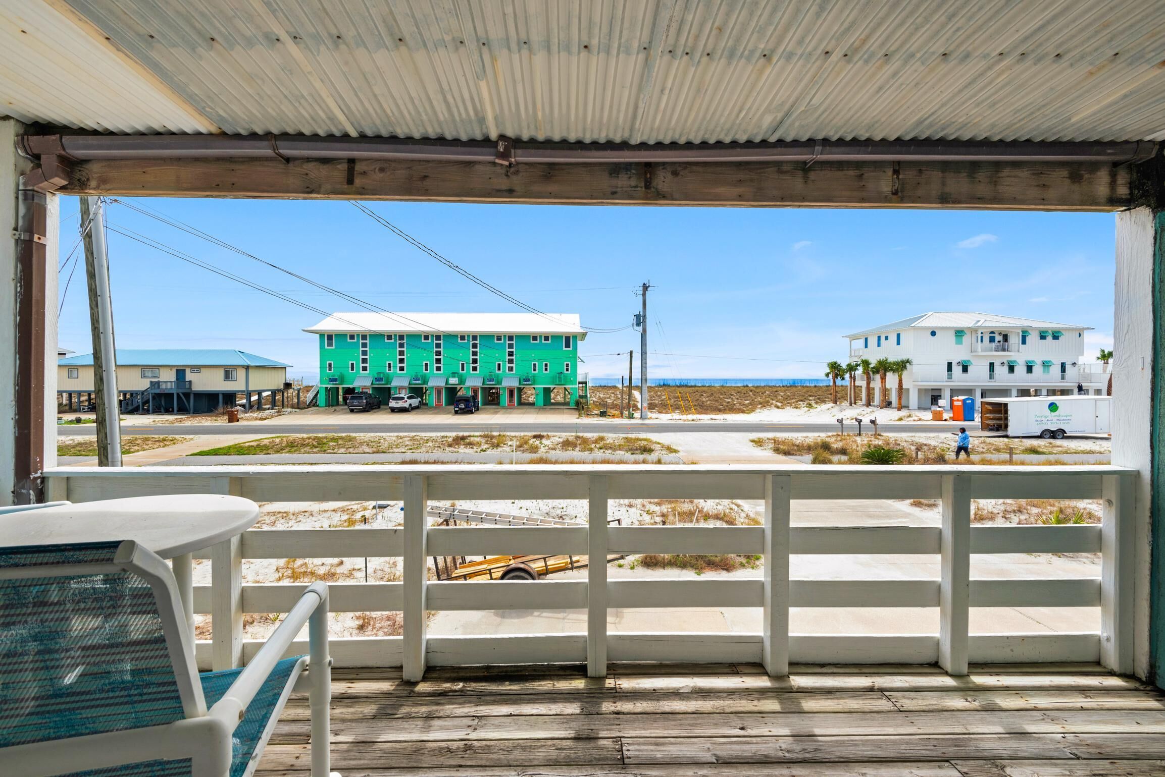 A beachside view from a covered porch with a white railing, overlooking a road with buildings, including a green house and a white house with palm trees.